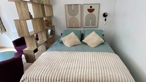 A bedroom with a wooden floor, a bed with a beige blanket, two pillows, a wooden side table, and a wall with two abstract art pieces.