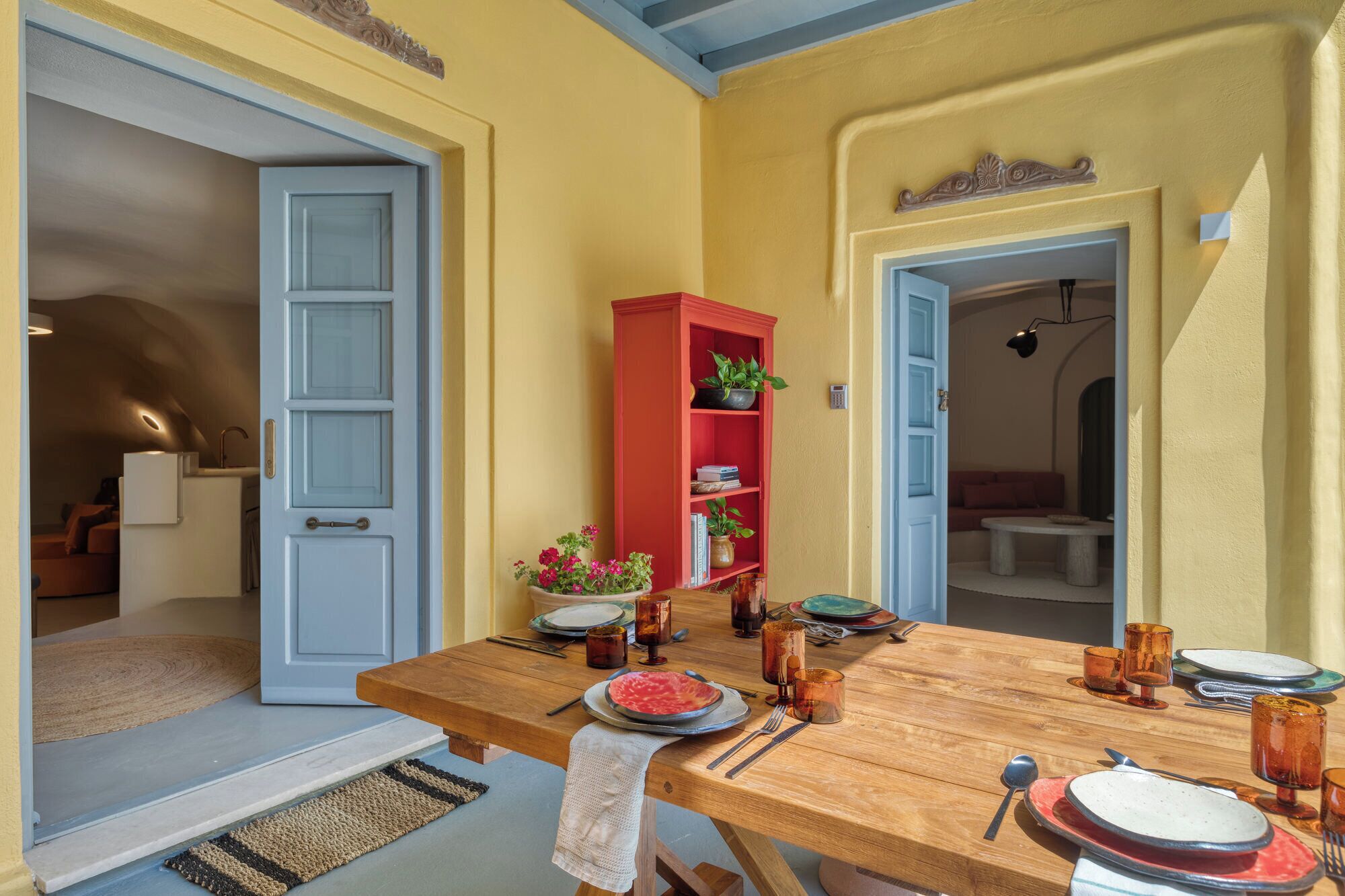 A dining area with a wooden table set for two, a red bookshelf, and a door leading to another room.