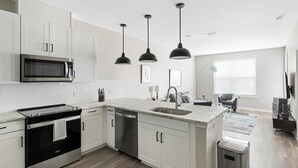 Una cocina moderna con gabinetes blancos, microondas integrado, estufa y lavavajillas. Hay lámparas colgantes sobre la isla y una ventana con persianas al fondo.
