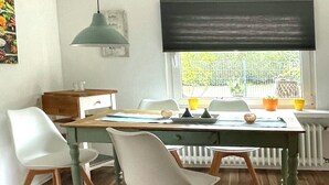 Un espace repas avec une table en bois, des chaises blanches, un luminaire suspendu vert et une fenêtre donnant sur un paysage verdoyant.