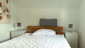 Une chambre à coucher avec une tête de lit en bois, un couvre-lit blanc à motifs, deux tables de chevet avec des lampes et une œuvre d’art accrochée au mur.