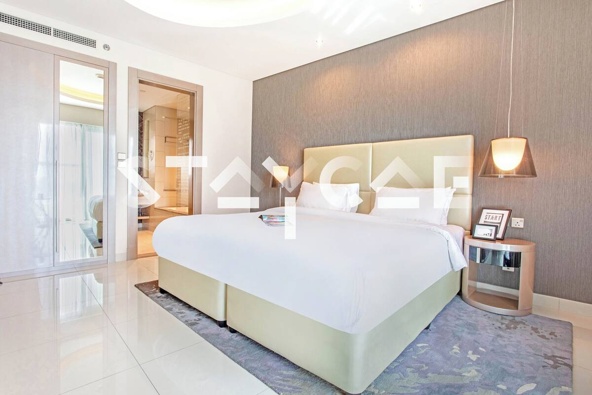 A modern hotel room with a large bed, a bedside table with a lamp, and a glass door leading to a bathroom.