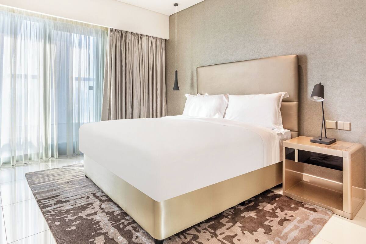 A modern hotel room with a large bed, a bedside table with a lamp, and a view of the city through sheer curtains.