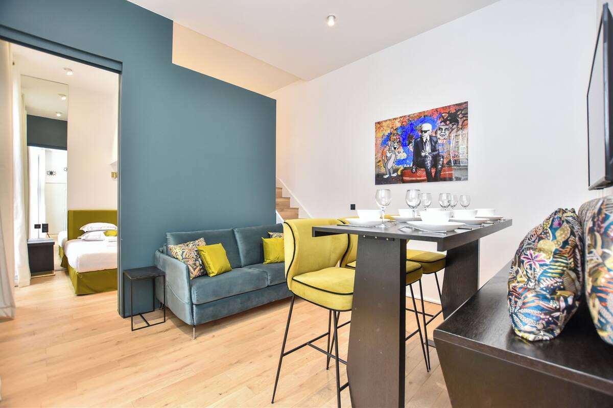 A modern room with a blue sofa, yellow chairs, a dining table with glasses, and a painting on the wall.