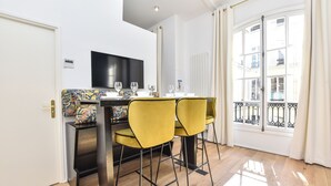 Un comedor con una mesa dispuesta para cuatro, sillas amarillas, un televisor negro en la pared y vistas de la ciudad a través de un ventanal grande.