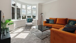Una sala de estar moderna con una mesa de centro de vidrio, un sofá naranja y un sillón blanco con un cojín azul.