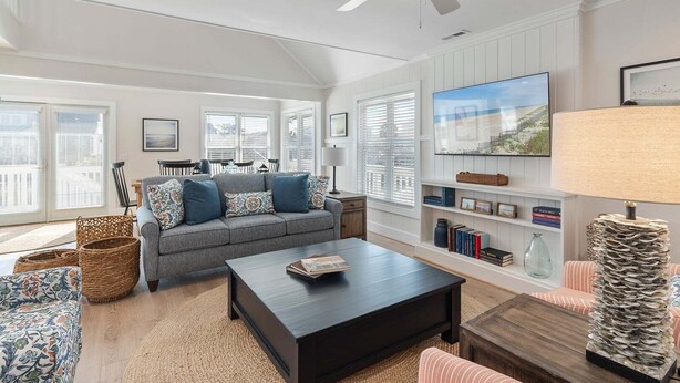 A spacious living room with a grey sofa, a coffee table, and a TV. There are wicker baskets, a bookshelf, and a lamp. - Relax in this charming home in Holden Beach featuring 3 bedrooms. (Holden Beach)