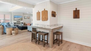 A kitchen with a white countertop, wooden stools, and a dining area with a sofa and a TV. - Relax in this charming home in Holden Beach featuring 3 bedrooms. (Holden Beach)