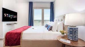 A hotel room with a bed, a TV mounted on the wall, a window with curtains, a bedside table with a lamp, and a small potted plant. - Staycae Holiday Homes - Suburbia (Dubai)