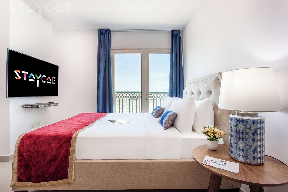 A hotel room with a bed, a TV mounted on the wall, a window with curtains, a bedside table with a lamp, and a small potted plant.