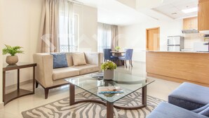 A modern living room with a glass coffee table, beige sofa, and a dining area with blue chairs. - Staycae Holiday Homes - Suburbia (Dubai)