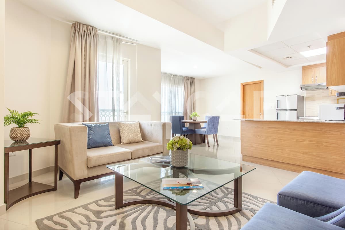 A modern living room with a glass coffee table, beige sofa, and a dining area with blue chairs.