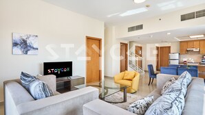 A modern living room with a sofa, armchair, and glass coffee table. A TV is on a stand, and there is a staircase leading to another level. - Staycae Holiday Homes - Suburbia (Dubai)