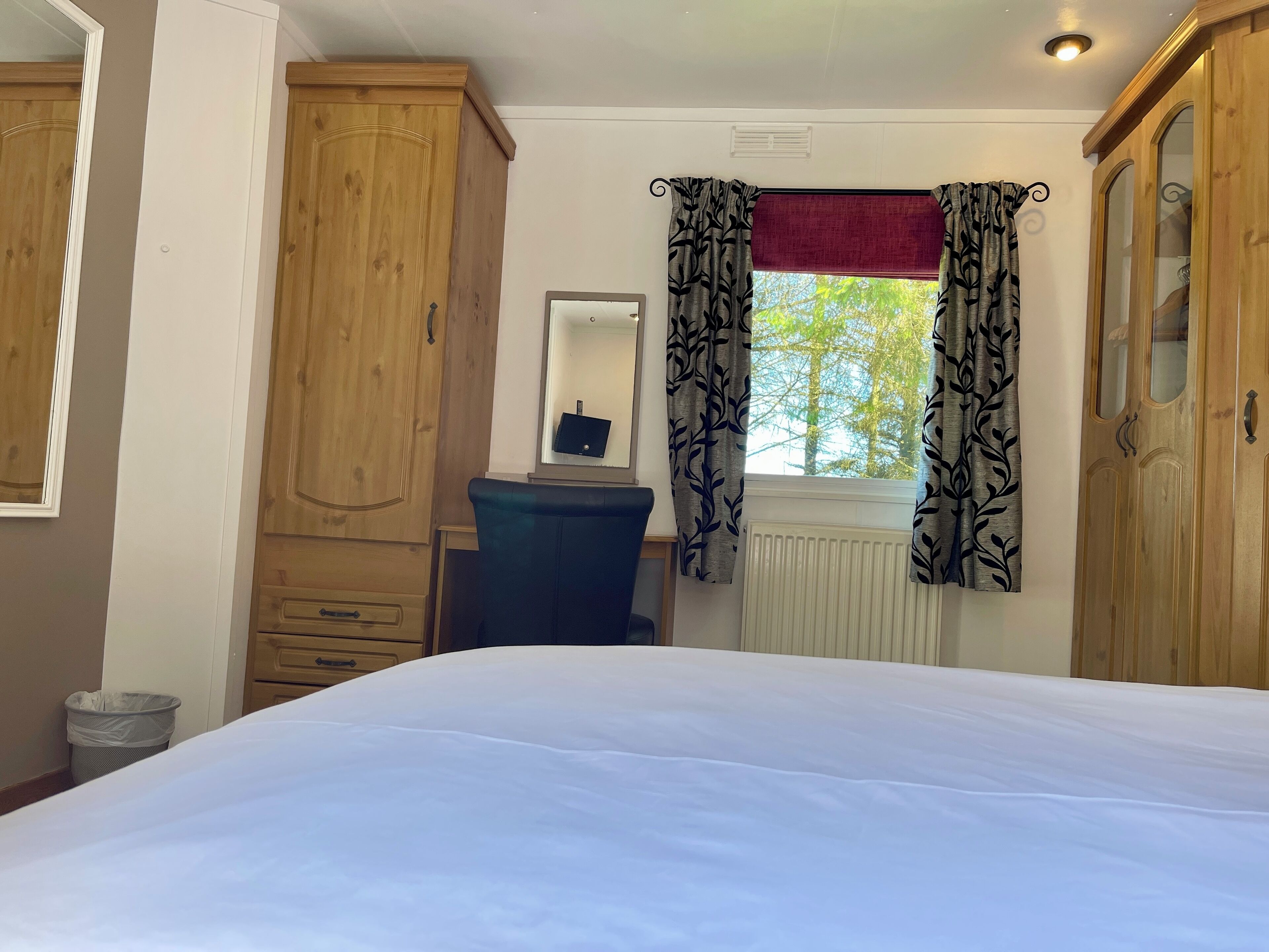 A bedroom with a bed, wooden wardrobe, a desk with a mirror, and a window with patterned curtains.