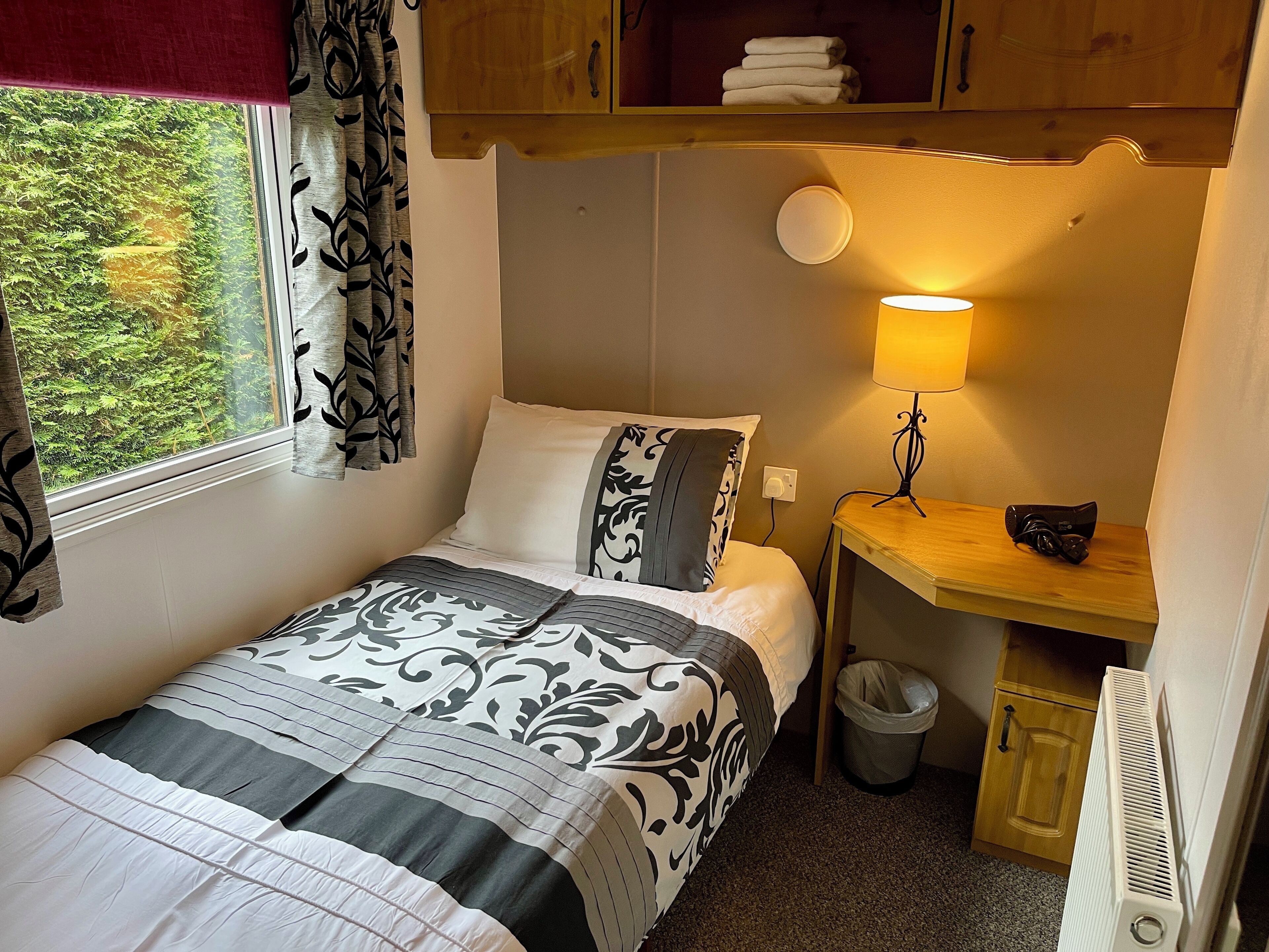 A compact bedroom with a single bed, a bedside table with a lamp, a small trash bin, and a radiator.