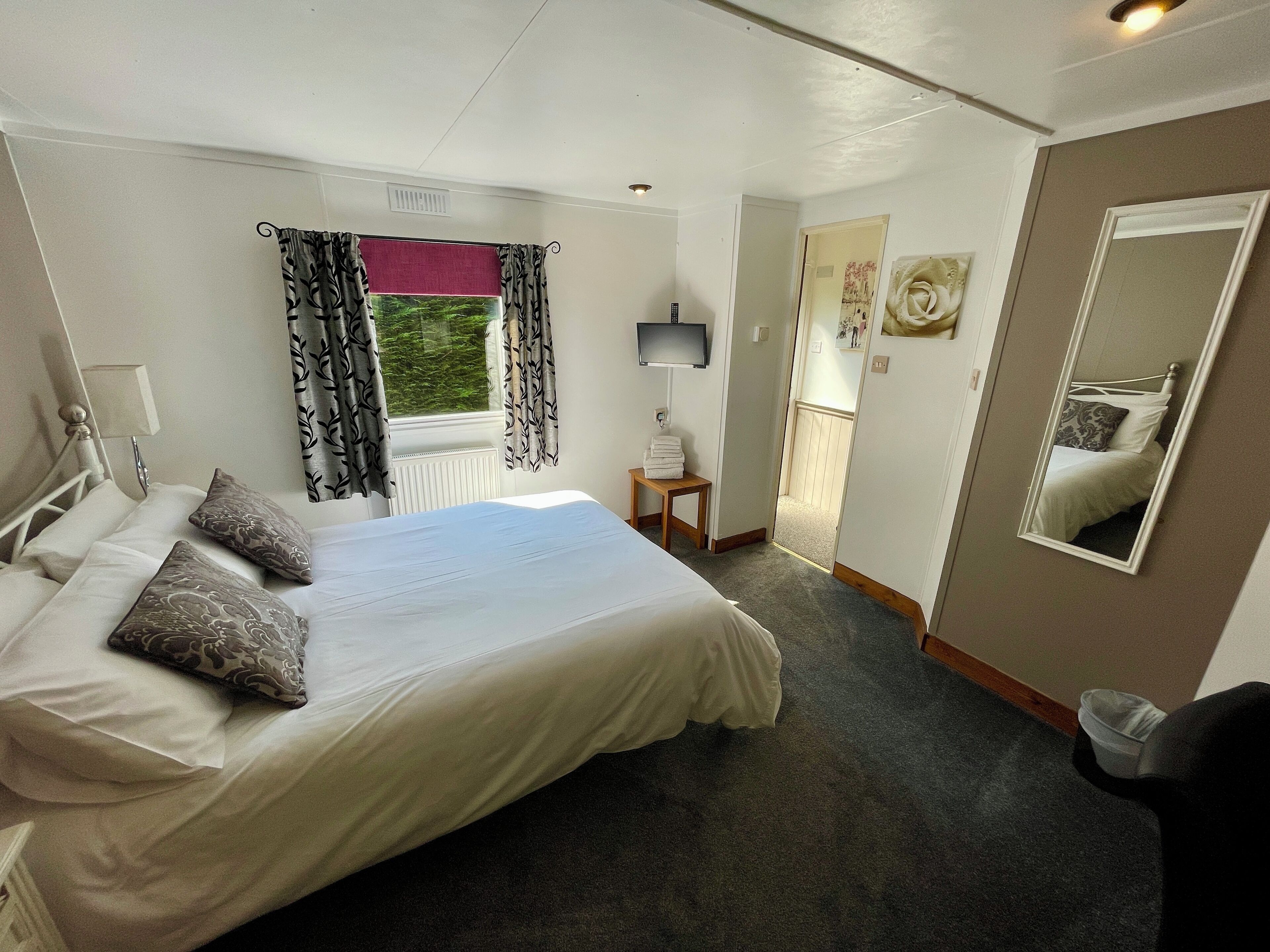A neatly arranged bedroom with a bed, pillows, a bedside table, a lamp, a window with curtains, a mirror, and a door leading to another room.