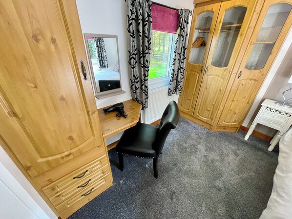 A bedroom with a wooden wardrobe, a desk with a chair, a mirror, and a window with curtains.