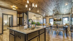 A spacious kitchen with granite countertops, a central island, and a stone backsplash. There is a dining area with wooden chairs and a table, and a living room with a sofa and a fireplace.