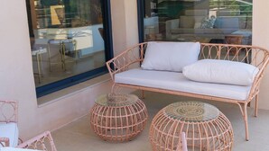 Un balcón con mobiliario de mimbre, incluyendo un sofá y dos mesas redondas, con una puerta de cristal que refleja el interior.