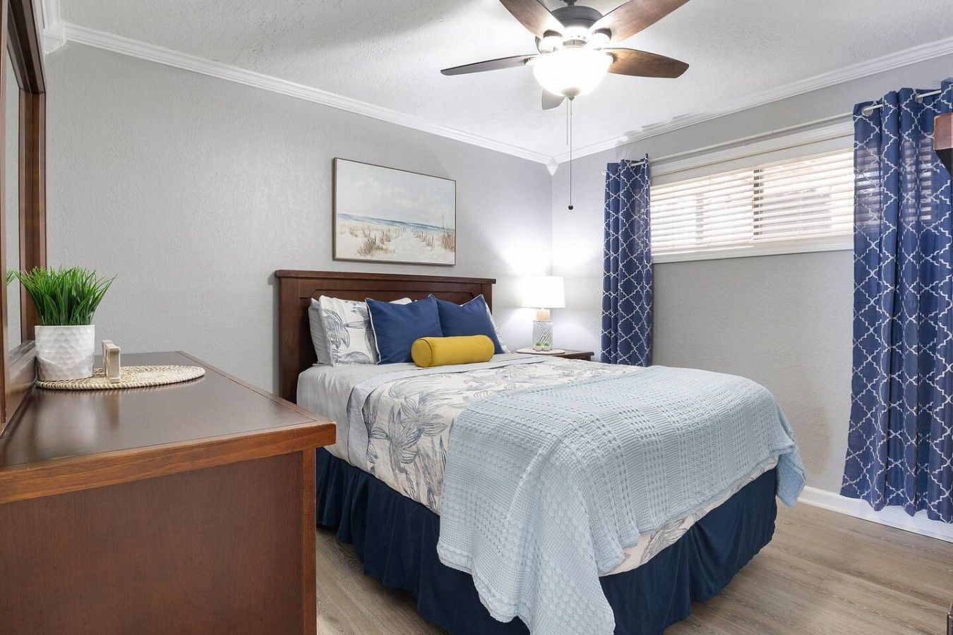 Une chambre à coucher comprenant un lit, une table de chevet, une lampe, un ventilateur de plafond, une fenêtre avec des rideaux et un tableau accroché au mur.
