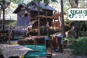 A miniature golf course with a rustic wooden clubhouse, a water hazard, and a sign reading 'Sugar Creek Mini Golf.' - Boho Flamingo-Pools, Hot Tub, Pet Friendly (Village of Four Seasons)