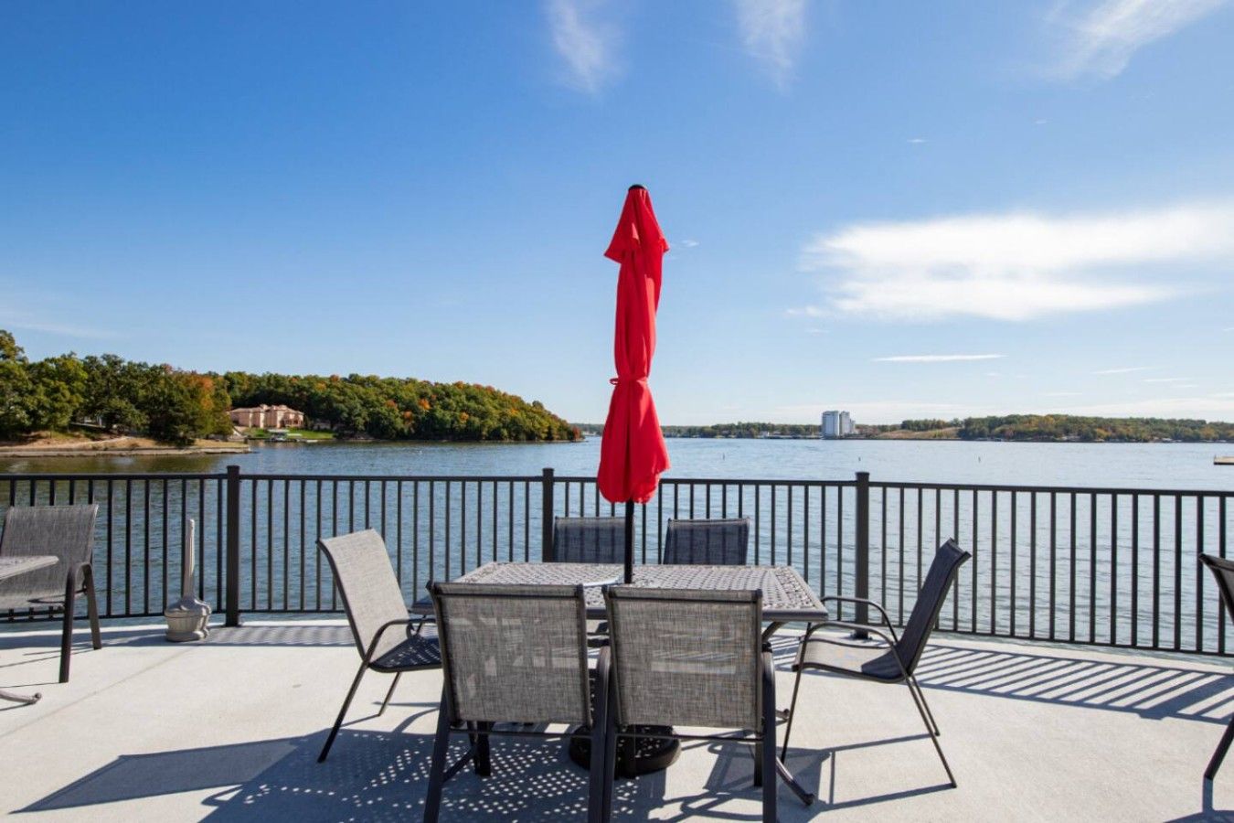 A deck with a table and chairs, a red umbrella, and a view of a lake with a distant building.