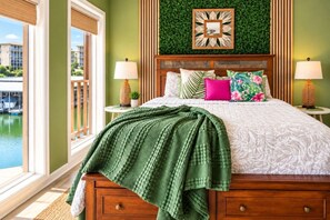 A bedroom with a bed, pillows, a green blanket, a wall-mounted clock, and a view of a body of water. - Boho Flamingo-Pools, Hot Tub, Pet Friendly (Village of Four Seasons)