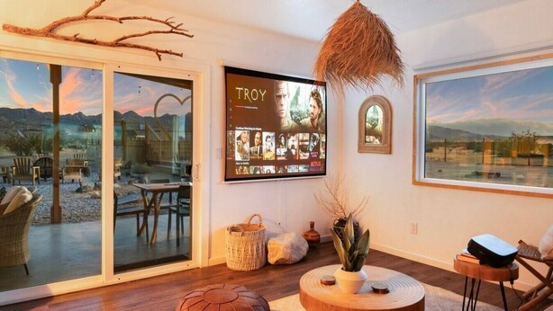 A modern living room with a large TV displaying a movie, a fireplace, and a view of a mountainous landscape. - The White Owl House ☆pool ☆private ☆gorgeous views (Joshua Tree)