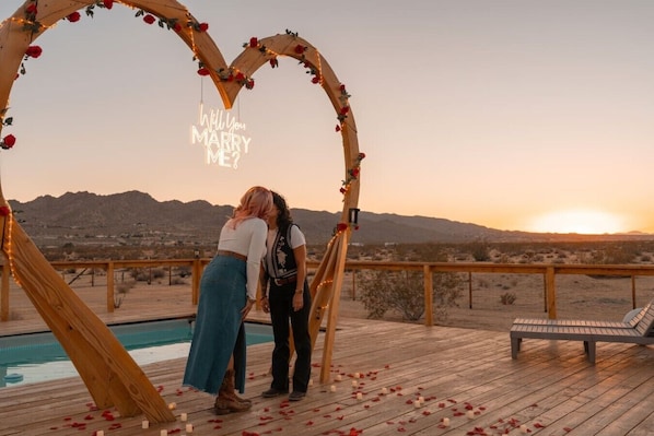 A wooden deck with a heart-shaped arch decorated with flowers and lights, a pool, and a sunset view.