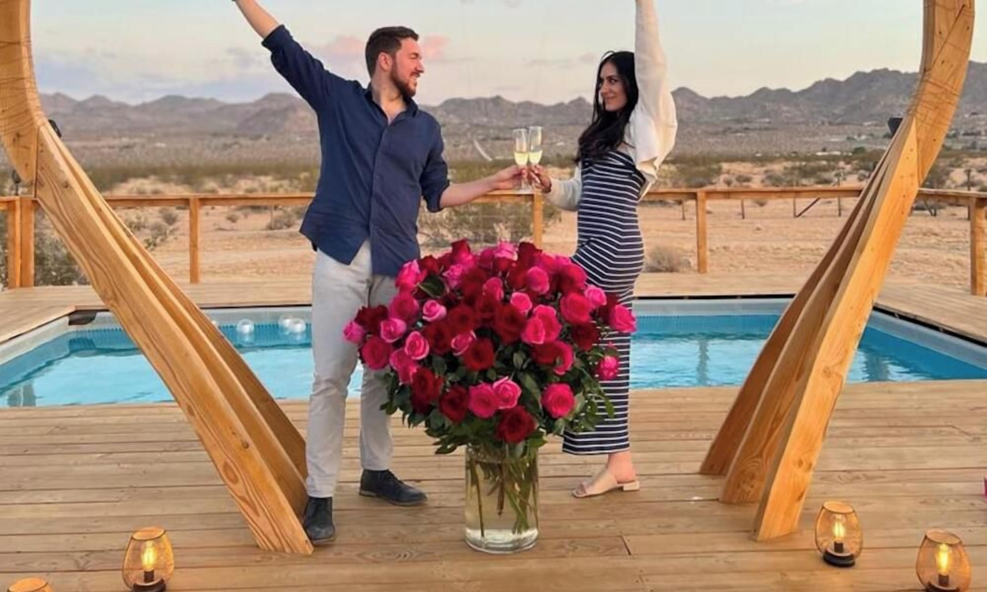 A wooden deck with a pool, a bouquet of red roses, and two people toasting with wine glasses.