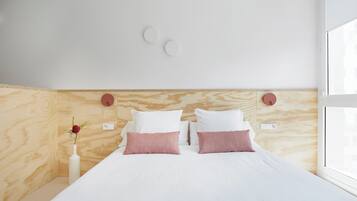 Una habitación de hotel moderna con cabecera de madera, una cama grande con ropa de cama blanca y rosa, un florero con una flor roja sobre una mesita de noche, y una ventana con vistas.