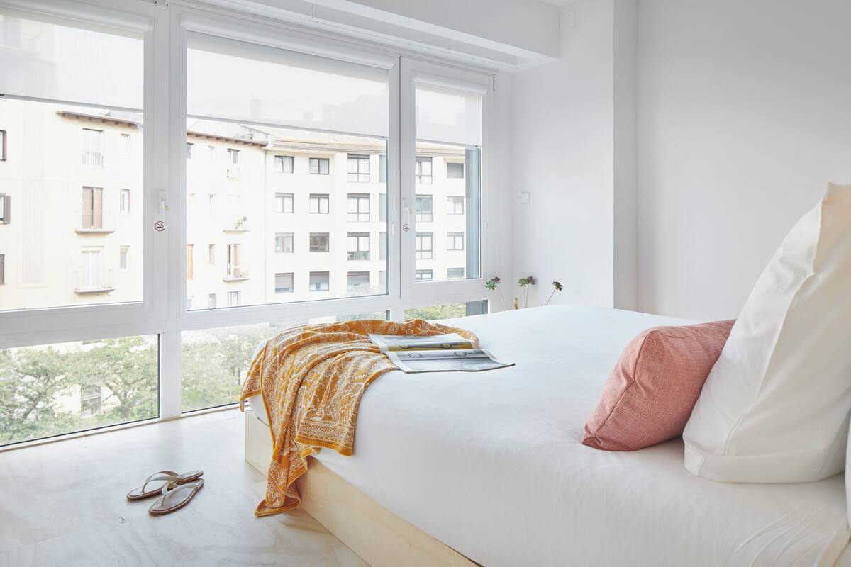A bedroom with a large window, a bed with white and pink pillows, and a yellow patterned throw blanket.