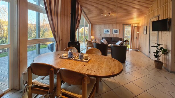 A cozy room with a wooden table, chairs, a sofa, and a TV. Large windows allow natural light and views of the outdoors.