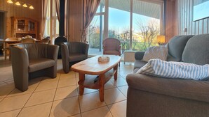A cozy living room with a fireplace, a dining area, and large windows offering a view of the outdoors.