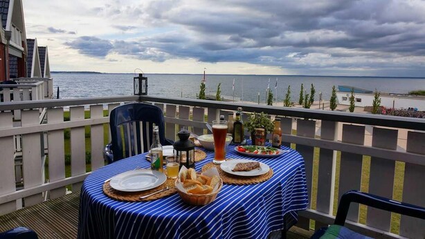 A dining table set on a balcony with a view of the sea, a bottle of wine, and a basket of bread.