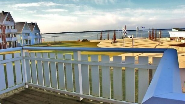 A coastal view with a wooden deck, a white fence, and a row of houses along the water.