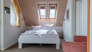 A bedroom with a sloped ceiling, two windows, a bed with white linens, a bedside table, and a pink chair.