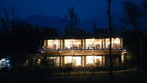 Un bâtiment à deux étages, doté de grandes fenêtres et d’un balcon, entouré d’arbres et avec une montagne en arrière-plan.
