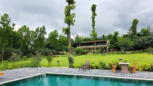 Une piscine dotée d’une terrasse en pierre, entourée d’une végétation luxuriante et avec un bâtiment visible au loin.