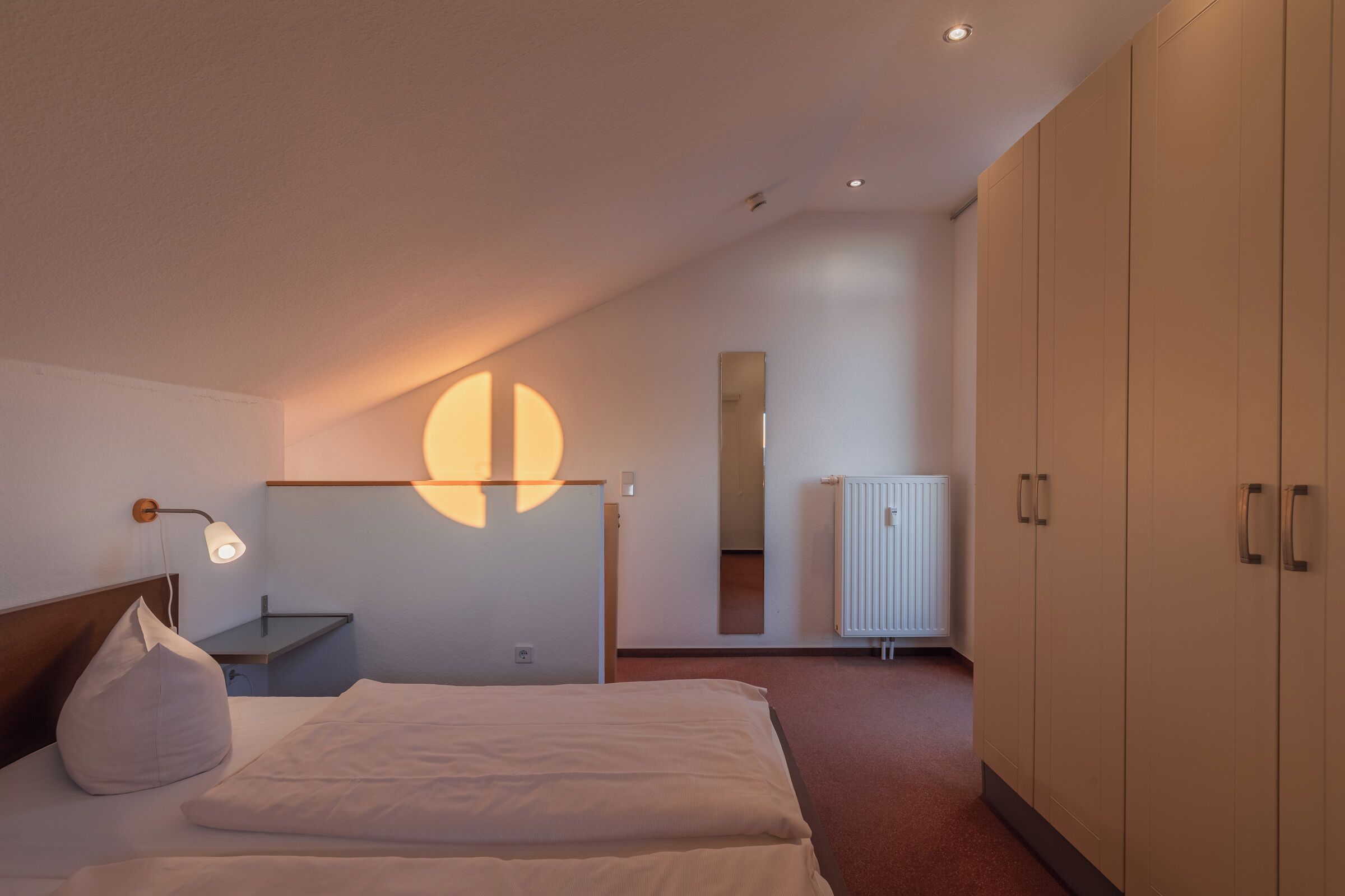 A modern hotel room with a bed, bedside table, lamp, wardrobe, and a large mirror.