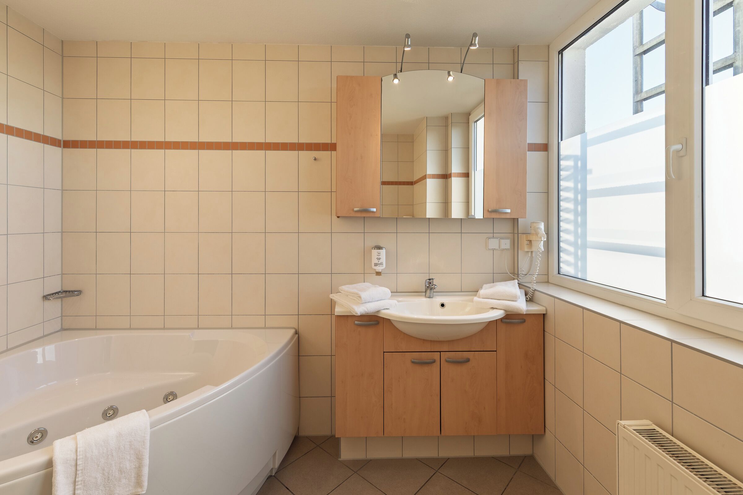 A modern bathroom with a bathtub, two sinks, a large mirror, and a window providing natural light.