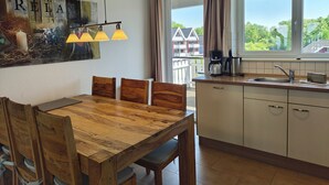 A dining area with a wooden table and chairs, a kitchen with a sink and cabinets, and a view of houses through a window.