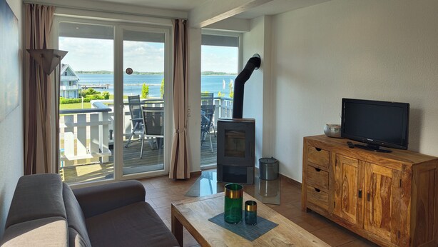 A living room with a sofa, a television on a wooden cabinet, a fireplace, and a sliding glass door leading to a balcony with a view of the water.