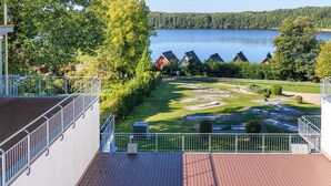 A view of a lakeside property with a wooden deck, a grassy area with a playground, and several houses with sloped roofs.