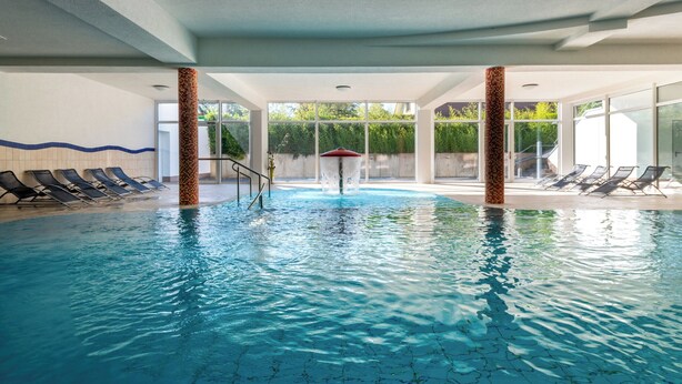 An indoor swimming pool with clear blue water, surrounded by lounge chairs and a glass wall.
