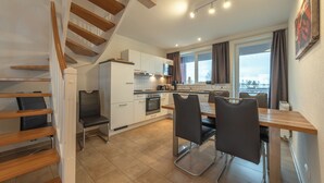 Una cocina moderna con armarios blancos, una zona de comedor con mesa de madera y sillas negras, y una escalera con escalones de madera.