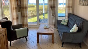 A living room with a sofa, armchair, and a small table. There are large windows with curtains, a view of the outdoors, and a painting on the wall.