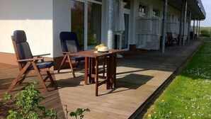 Area esterna coperta con mobili in legno e un tavolo, adiacente a un edificio con ampie finestre.