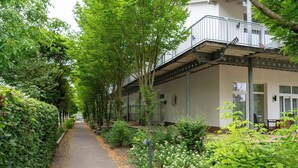 Un sentiero fiancheggiato da vegetazione rigogliosa e fiori bianchi, che conduce a un edificio con un balcone e un portico coperto.
