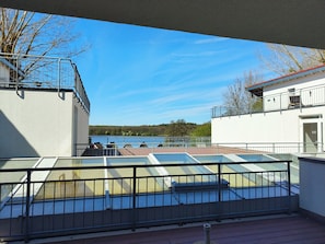 A rooftop terrace with a glass balustrade, a view of a lake, and a building with a white facade and red roof.